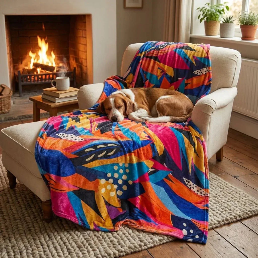 Un chien aux poils marron et blanc est installĂ© sur un fauteuil couvert dâun plaid Panache aux couleurs jaune, orange, bleu, violet et fuchsia , Ă cĂŽtĂ© du feu de cheminĂ©e.