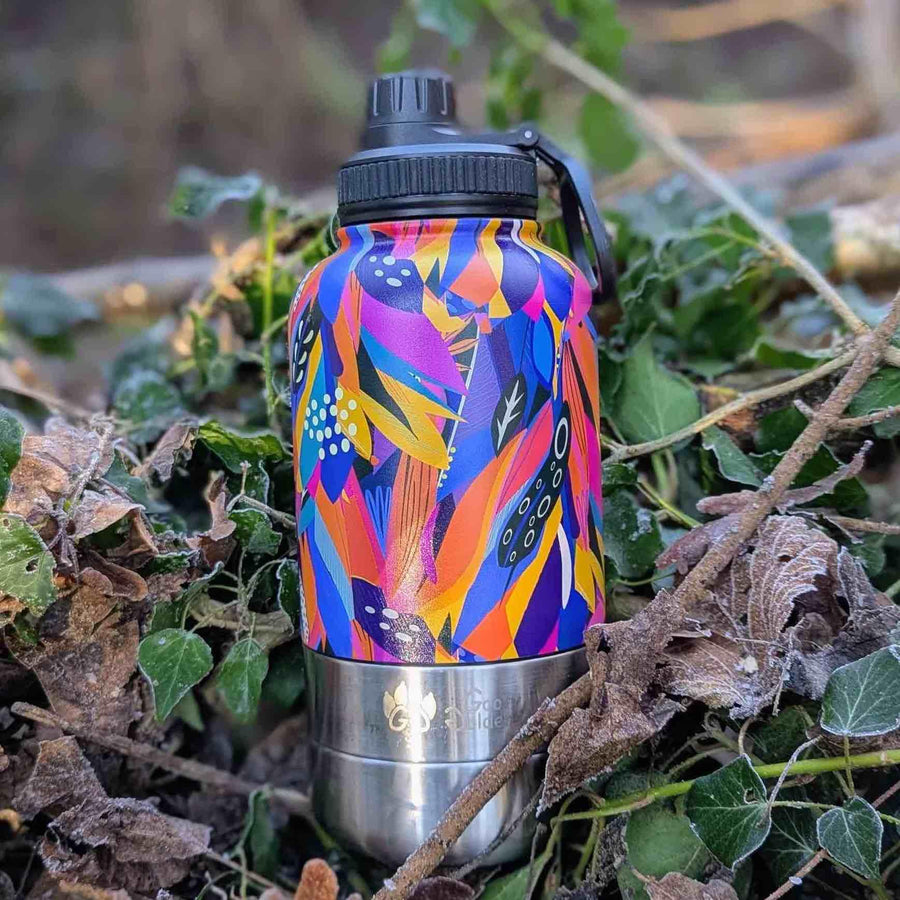 Gourde de randonnée 3-en-1 pour chien et humain Goofy Goldens, aux motifs abstraits de plumes jaune, orange, bleu, violet et fuchsia, posée sur un sol couvert de feuilles.