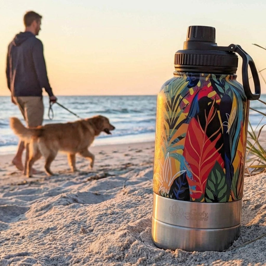 Au premier plan se trouve une gourde 3-en-1 de la gamme Tropical posée sur le sable, tandis qu’au loin un maître marche avec son chien au bord de la mer.