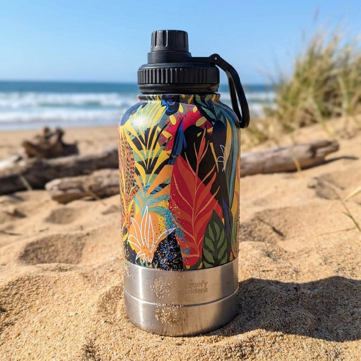 Une gourde 3-en-1 Tropical, décorée de motifs tropicaux jaunes, rouges, verts, oranges et noirs, est posée sur le sable au bord de l'eau.