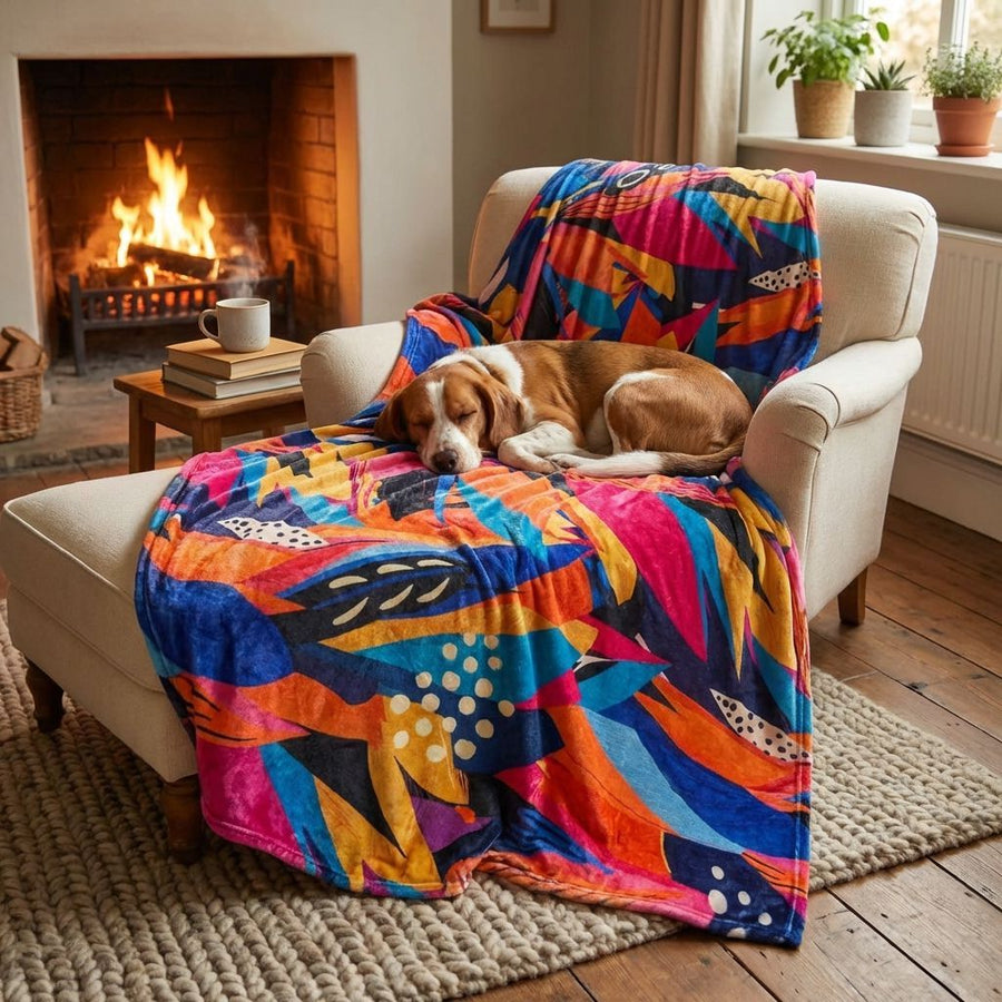 Un chien aux poils marron et blanc est installé sur un fauteuil couvert d’un plaid Panache aux couleurs jaune, orange, bleu, violet et fuchsia , à côté du feu de cheminée.